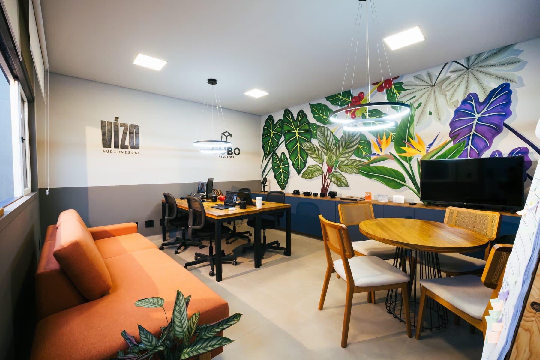 Sala do estúdio da Vízo com sofá laranja, mesa de trabalho com computadores, mesa redonda de madeira e um mural de folhagens na parede.