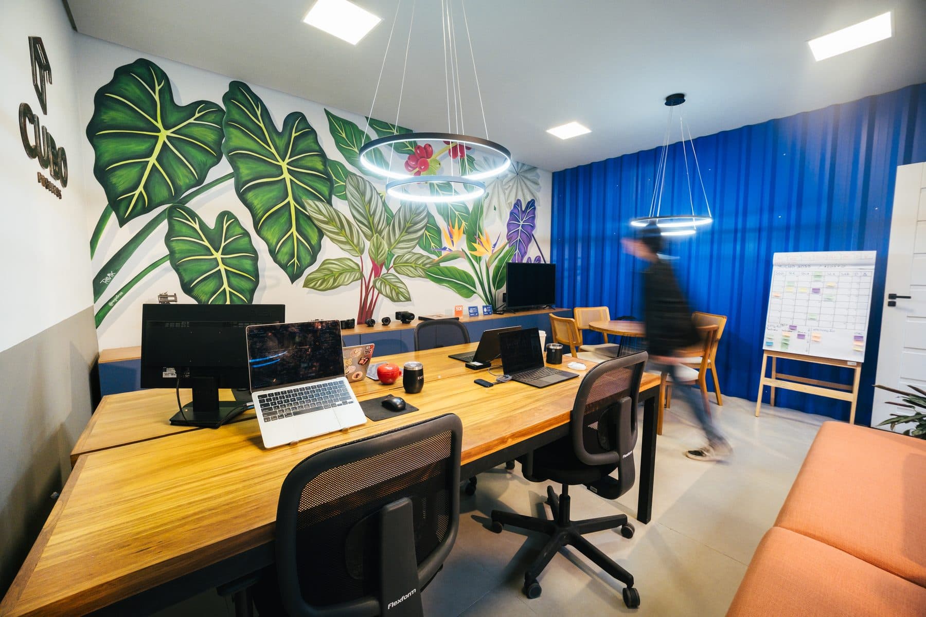 Mesa de trabalho com notebooks e monitores, mural de folhagens à esquerda, parede azul e quadro de planejamento ao fundo.