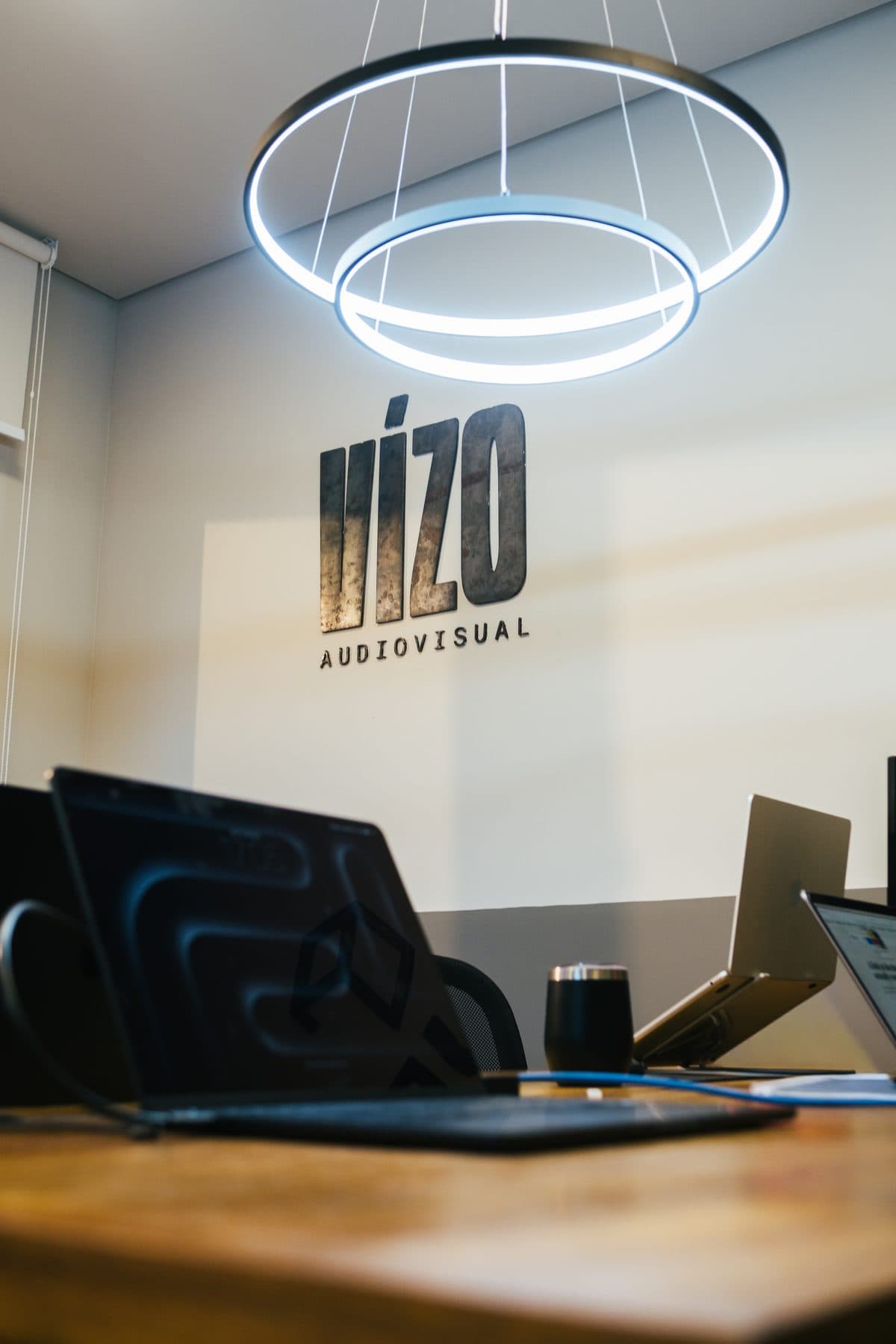 Letreiro metálico Vízo Audiovisual na parede, sob um lustre circular de luz, com notebooks sobre a mesa em primeiro plano.