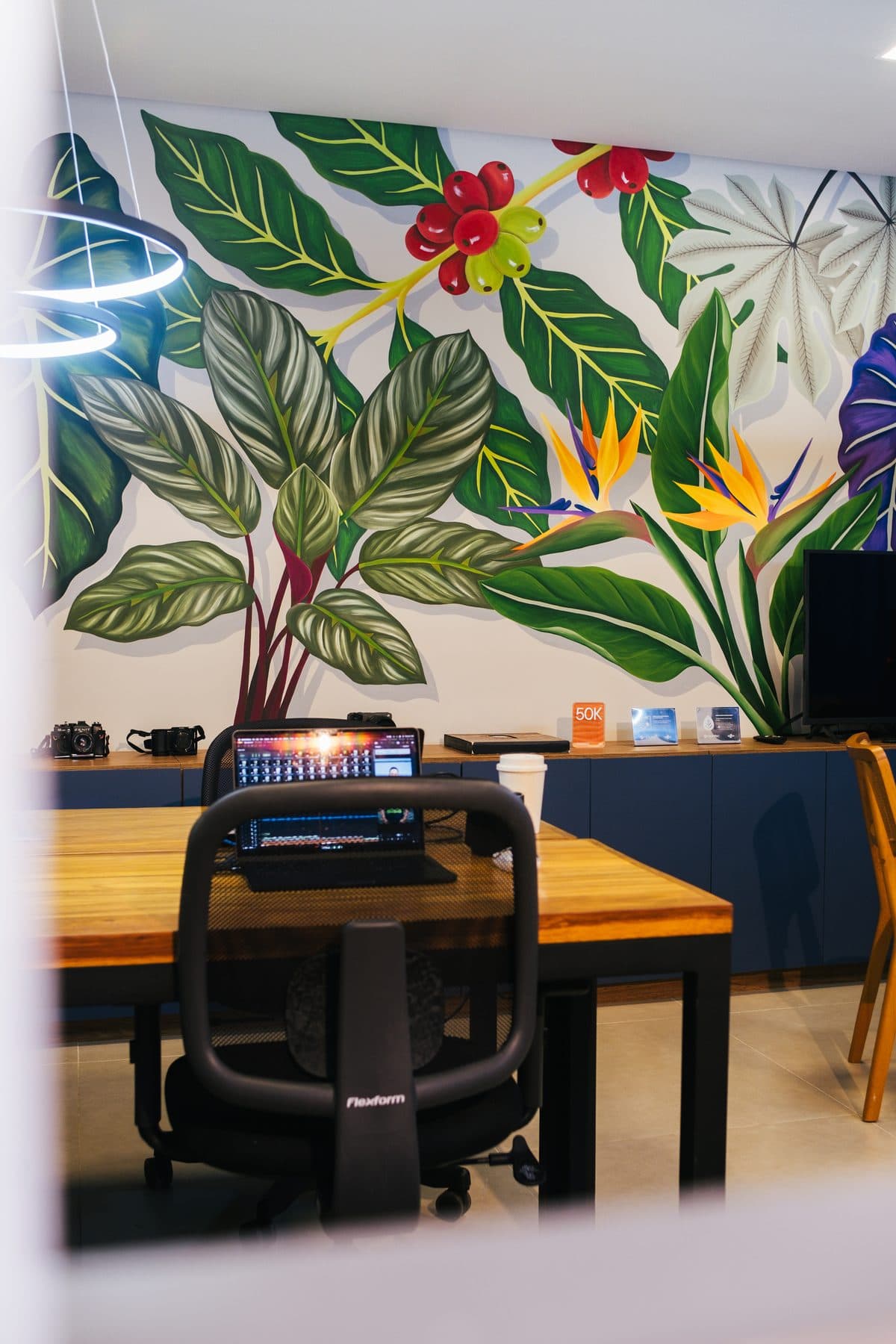 Mural de folhagens e flores do estúdio, com uma mesa de trabalho e um notebook de edição de vídeo em frente.
