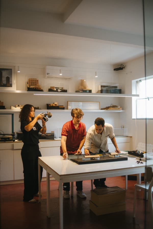 Equipe da Vízo gravando uma maquete de arquitetura sobre a mesa, vista através de uma janela de vidro, com outras maquetes nas prateleiras ao fundo.