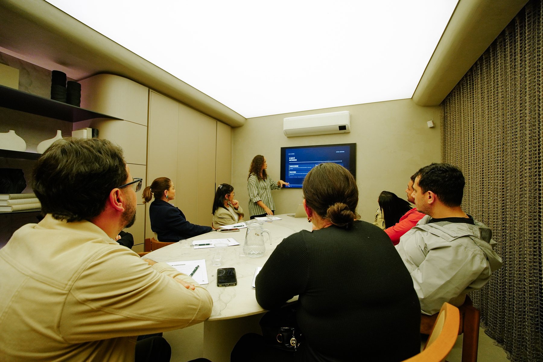 Sala de workshop com a facilitadora apresentando na TV enquanto os participantes acompanham em volta da mesa.