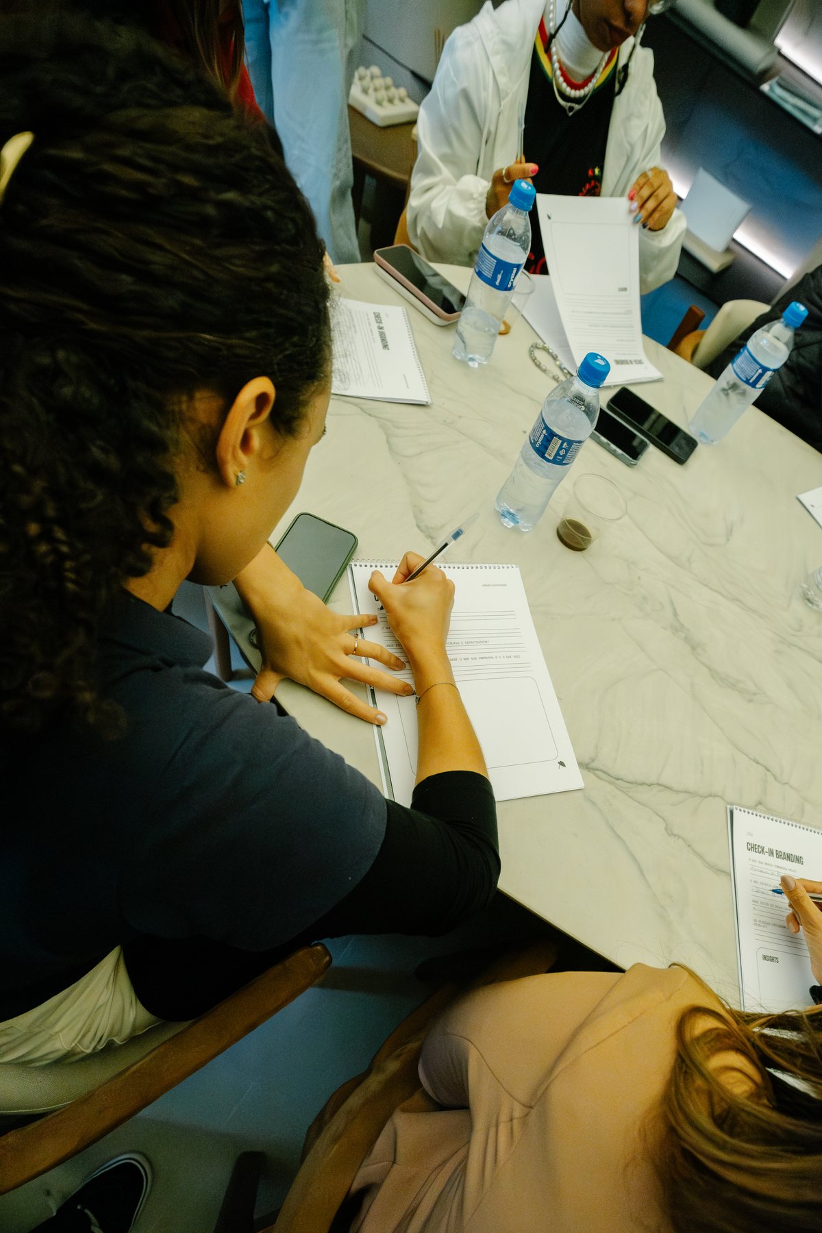 Vista de cima de participantes preenchendo o caderno “Check-in Branding” sobre a mesa.